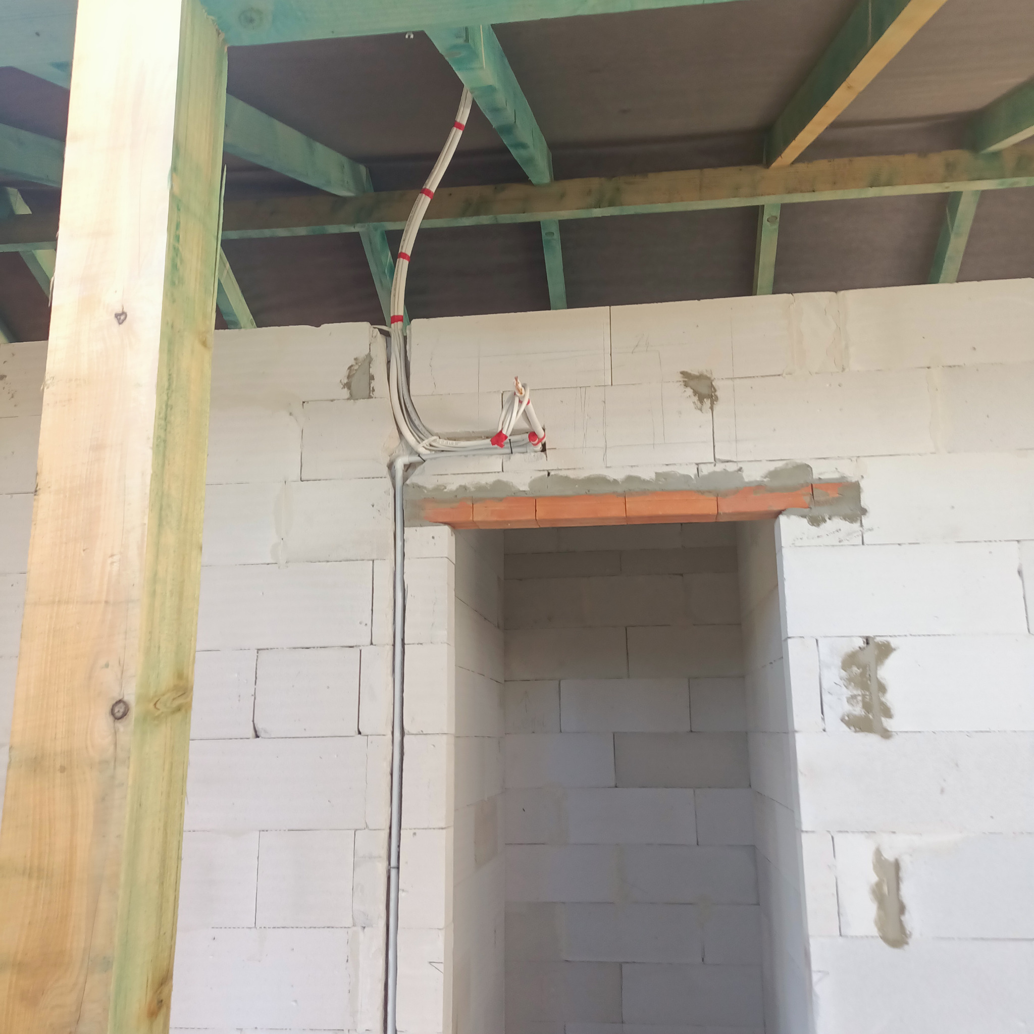 Wnętrze w stanie surowym: ściany z białych bloczków, drewniana konstrukcja stropu z widocznymi przewodami elektrycznymi zabezpieczonymi czerwonymi opaskami, otwór drzwiowy z nadprożem z czerwonej...