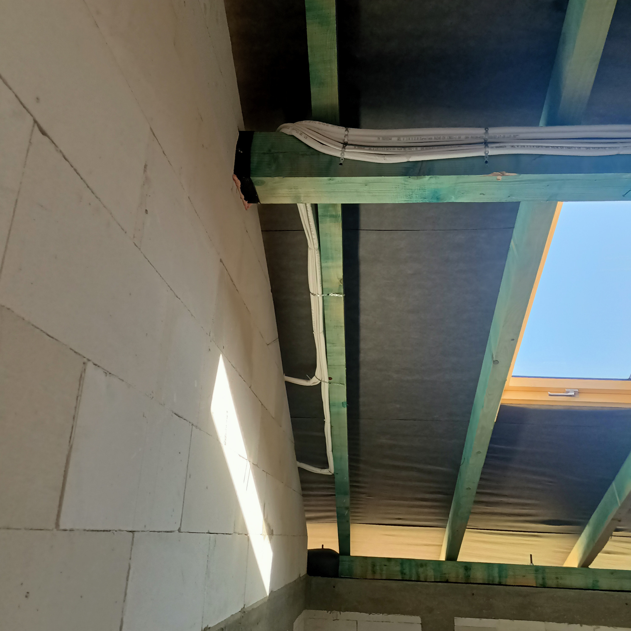 Wnętrze w stanie surowym: białe ściany z betonu komórkowego, zielone drewniane belki stropowe z czarną membraną, oraz białe przewody elektryczne przymocowane do belek. W tle widoczne okno dachowe...