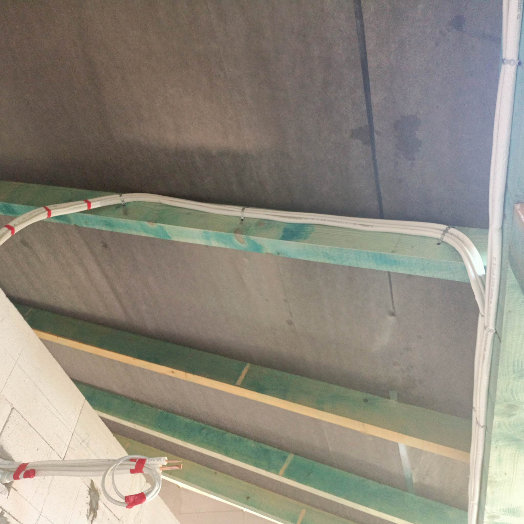 Białe przewody elektryczne zamocowane do drewnianej konstrukcji dachu z szarą membraną, instalacja w trakcie budowy, widoczne mocowania i oznaczenia na przewodach.