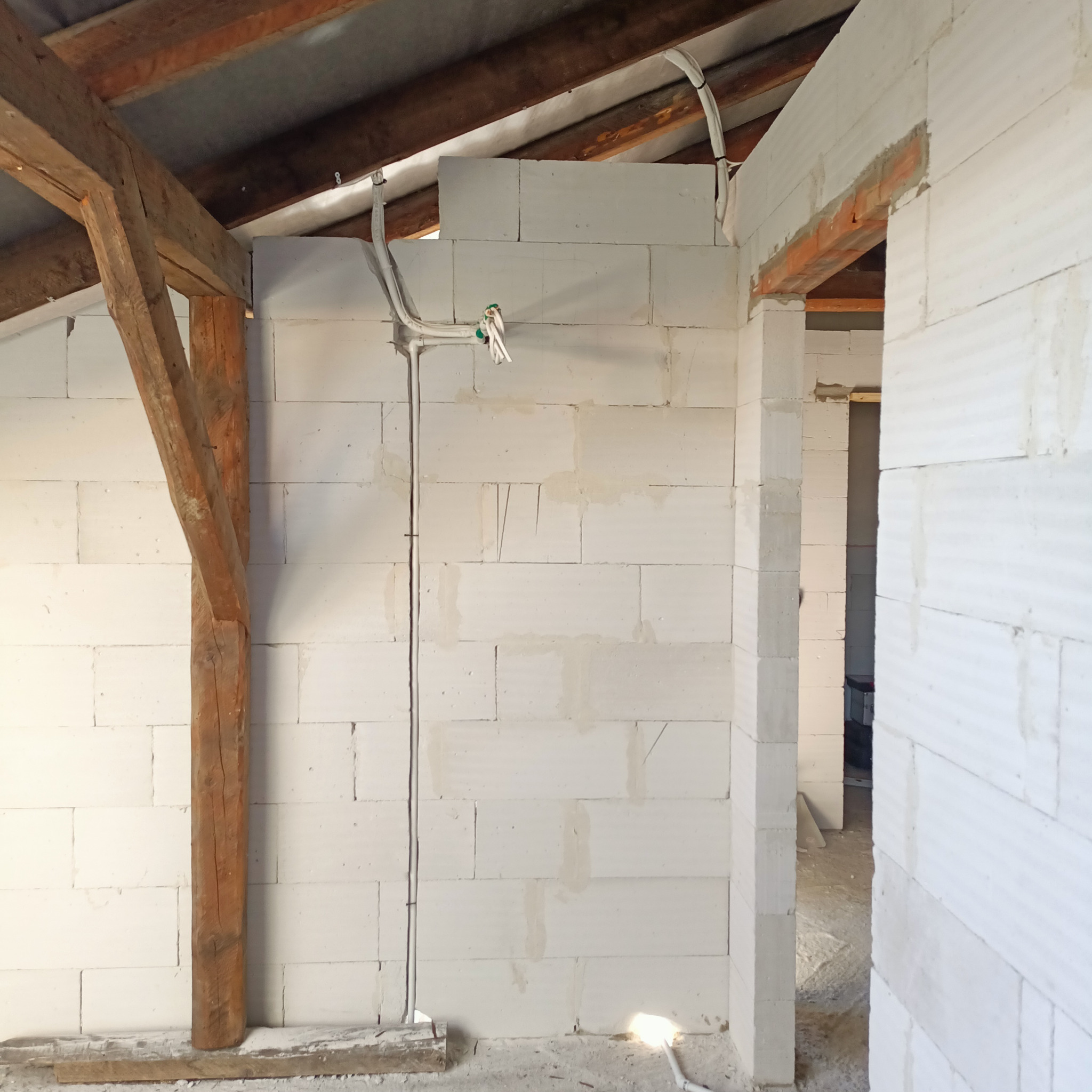 Wnętrze w trakcie budowy: ściany z białych bloczków, drewniana konstrukcja dachu, poprowadzone kable elektryczne z uchwytami, widoczne ślady zaprawy.