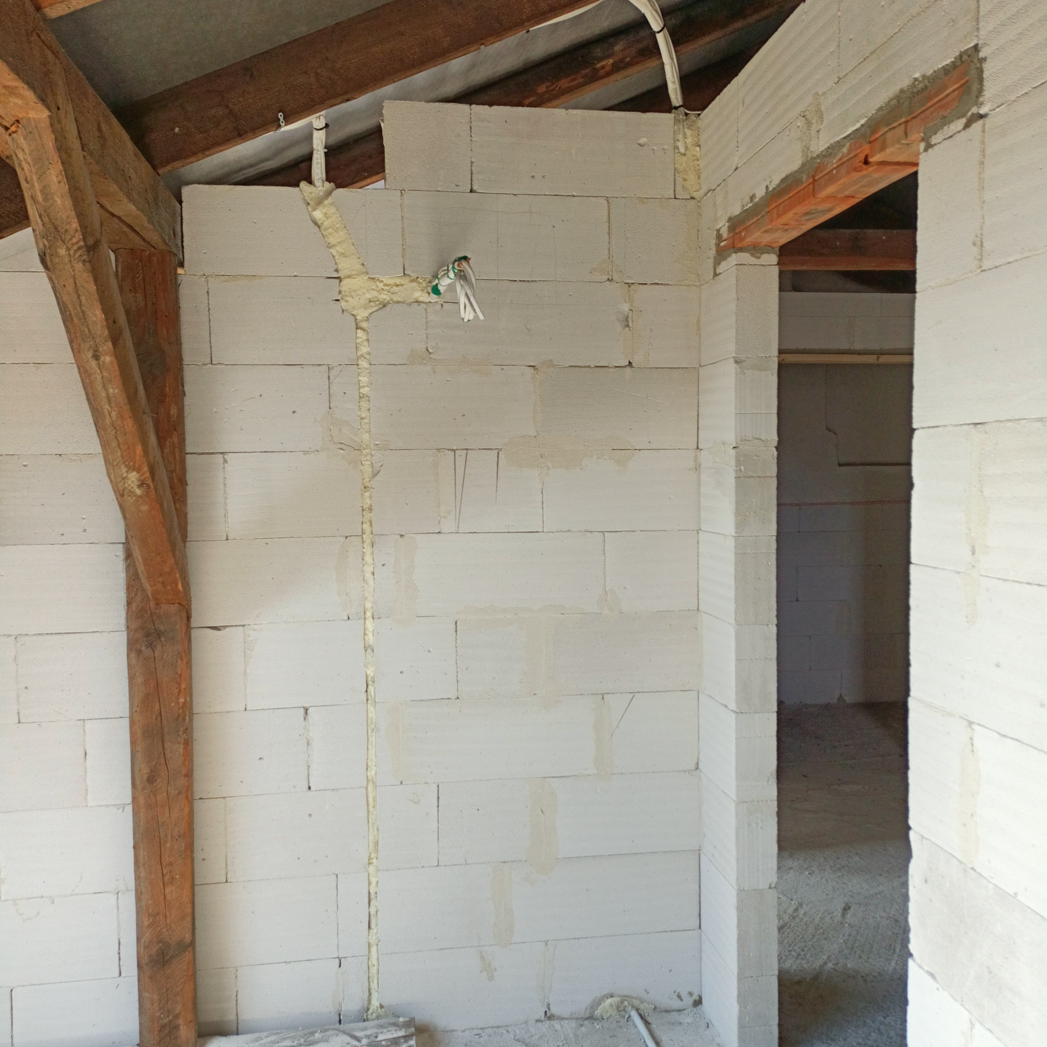 Wnętrze w trakcie budowy: ściany z białych bloczków, widoczne drewniane elementy konstrukcyjne, pionowy kanał wypełniony pianką montażową z wystającymi przewodami elektrycznymi.