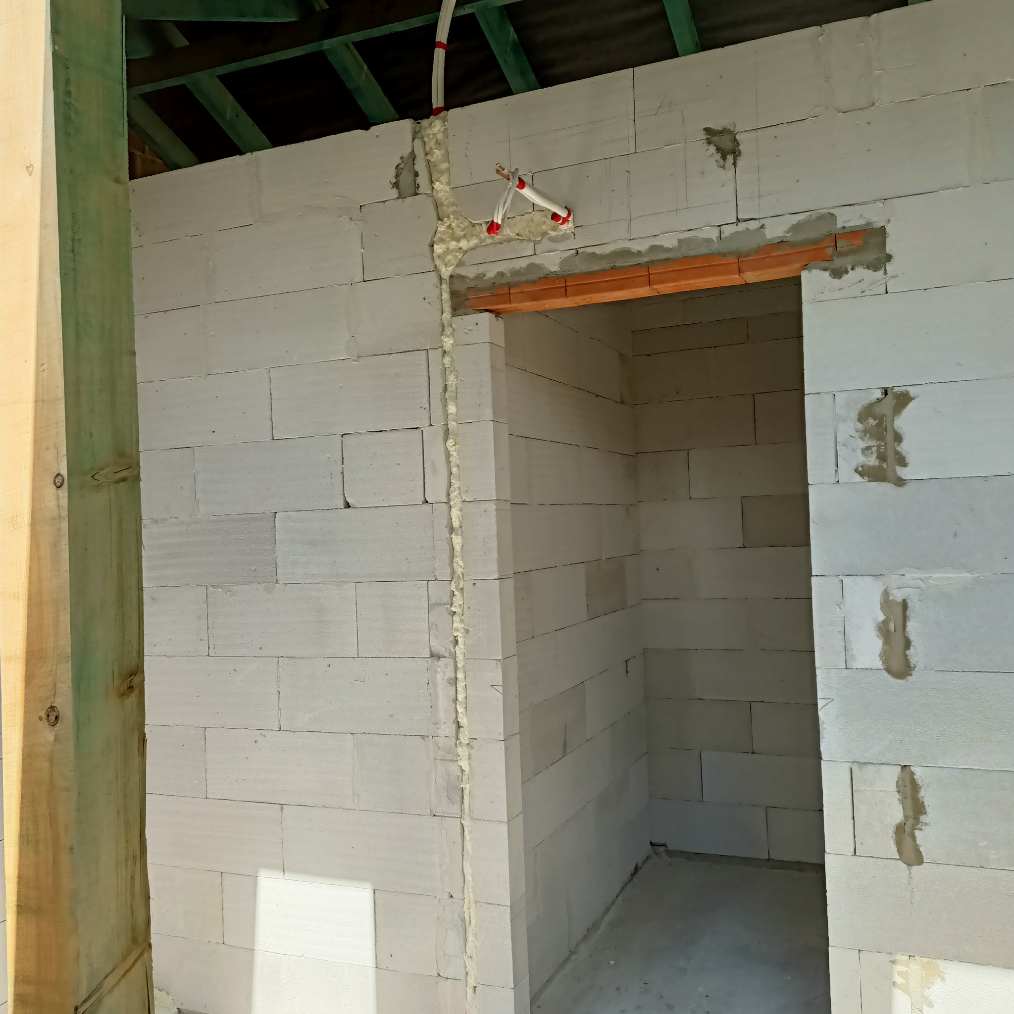 Wnętrze w trakcie budowy z białych bloczków, widoczne przewody elektryczne zabezpieczone pianką montażową, drewniana rama drzwiowa i zielona konstrukcja stropu.