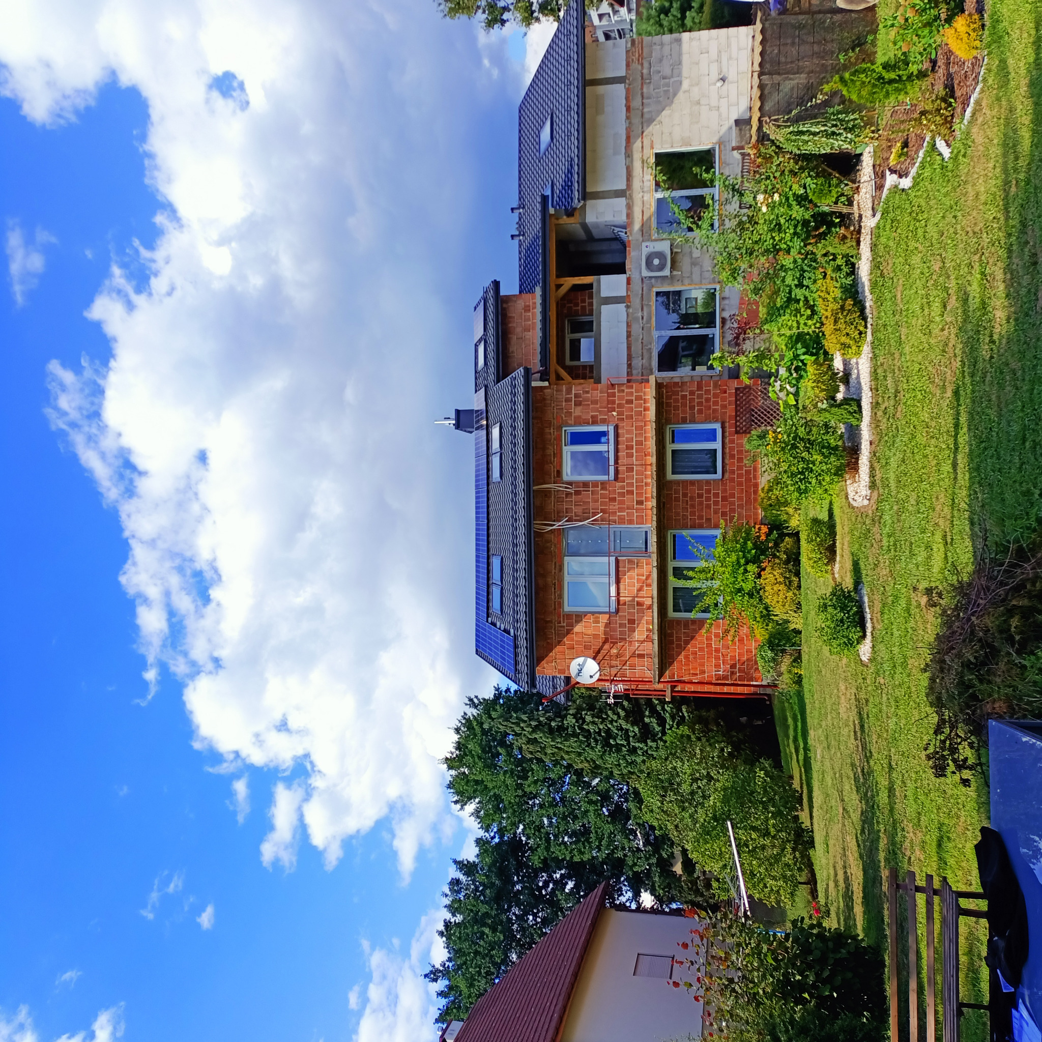 Dom w trakcie budowy z zamontowanymi panelami słonecznymi na dachu, widoczny klimatyzator na elewacji i bujna roślinność w ogrodzie.