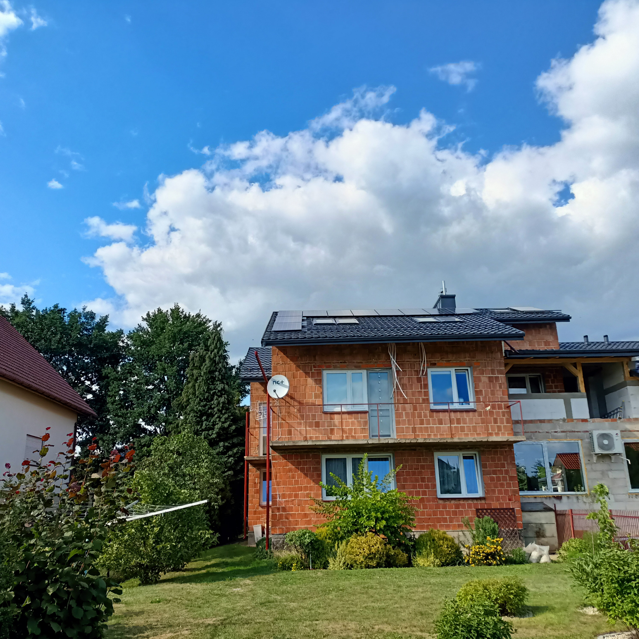Niedokończony dom z czerwonej cegły z panelami fotowoltaicznymi na dachu i anteną satelitarną na ścianie, widok z ogrodu w słoneczny dzień.