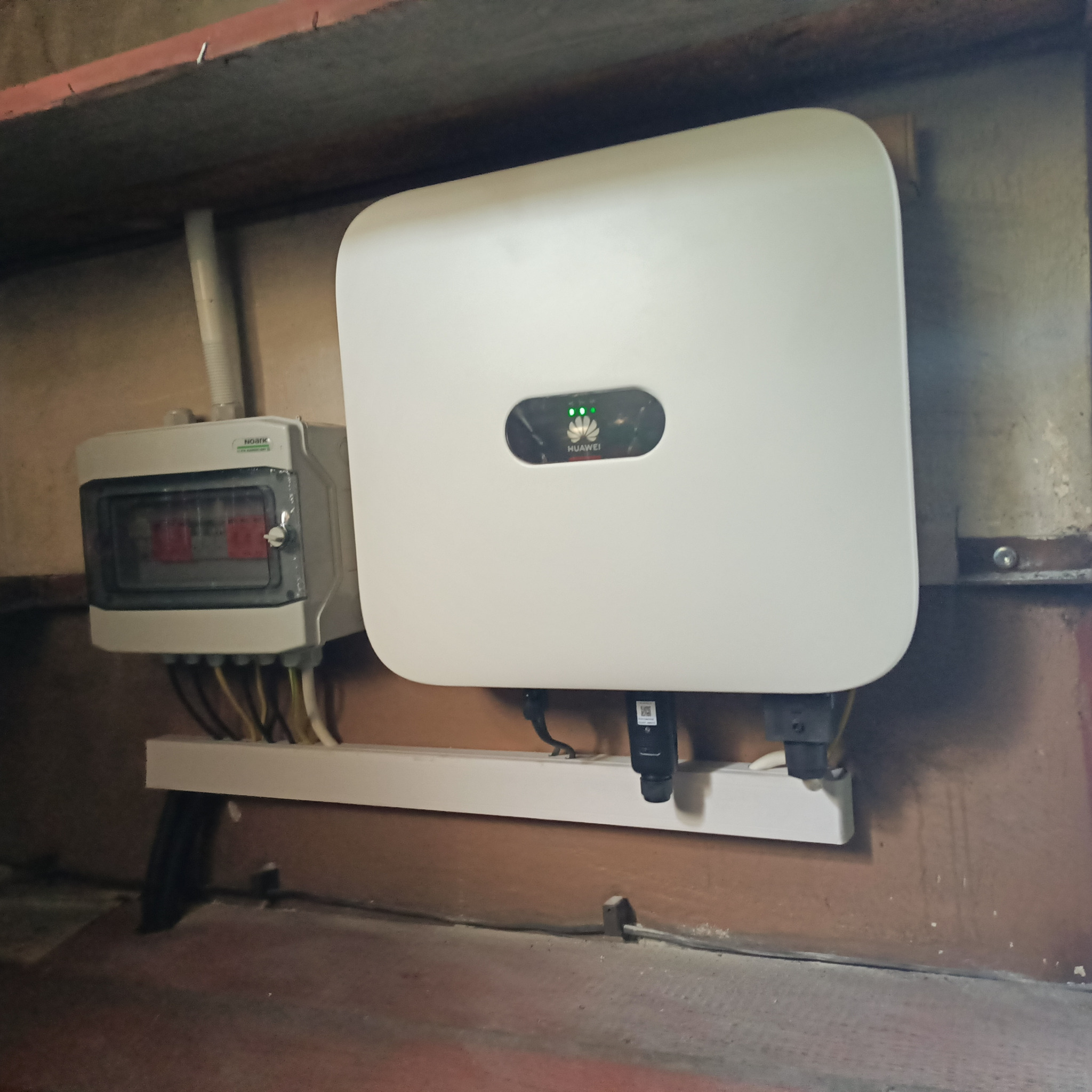 Biały inwerter Huawei z podłączonymi przewodami i obok skrzynka elektryczna z bezpiecznikami, zamontowane na ścianie wewnątrz pomieszczenia.