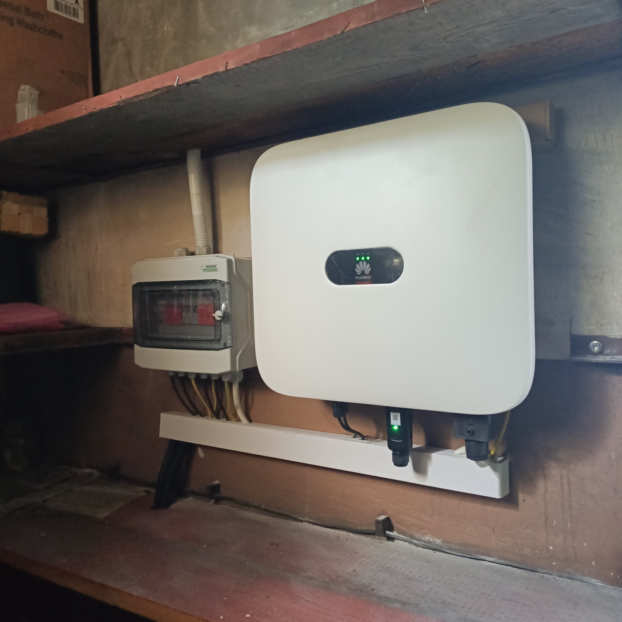 Biały falownik Huawei z zielonymi diodami LED i skrzynka bezpiecznikowa zamontowane na ścianie wewnątrz budynku, widoczne okablowanie.