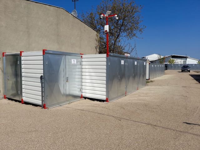 Rząd zewnętrznych, metalowych boksów magazynowych z numeracją, zabezpieczonych systemem monitoringu na czerwonym słupie, na żwirowym podłożu.