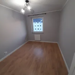 Pomalowane na szaro puste pomieszczenie z panelami podłogowymi, nowoczesną lampą sufitową i oknem z widokiem na budynek.