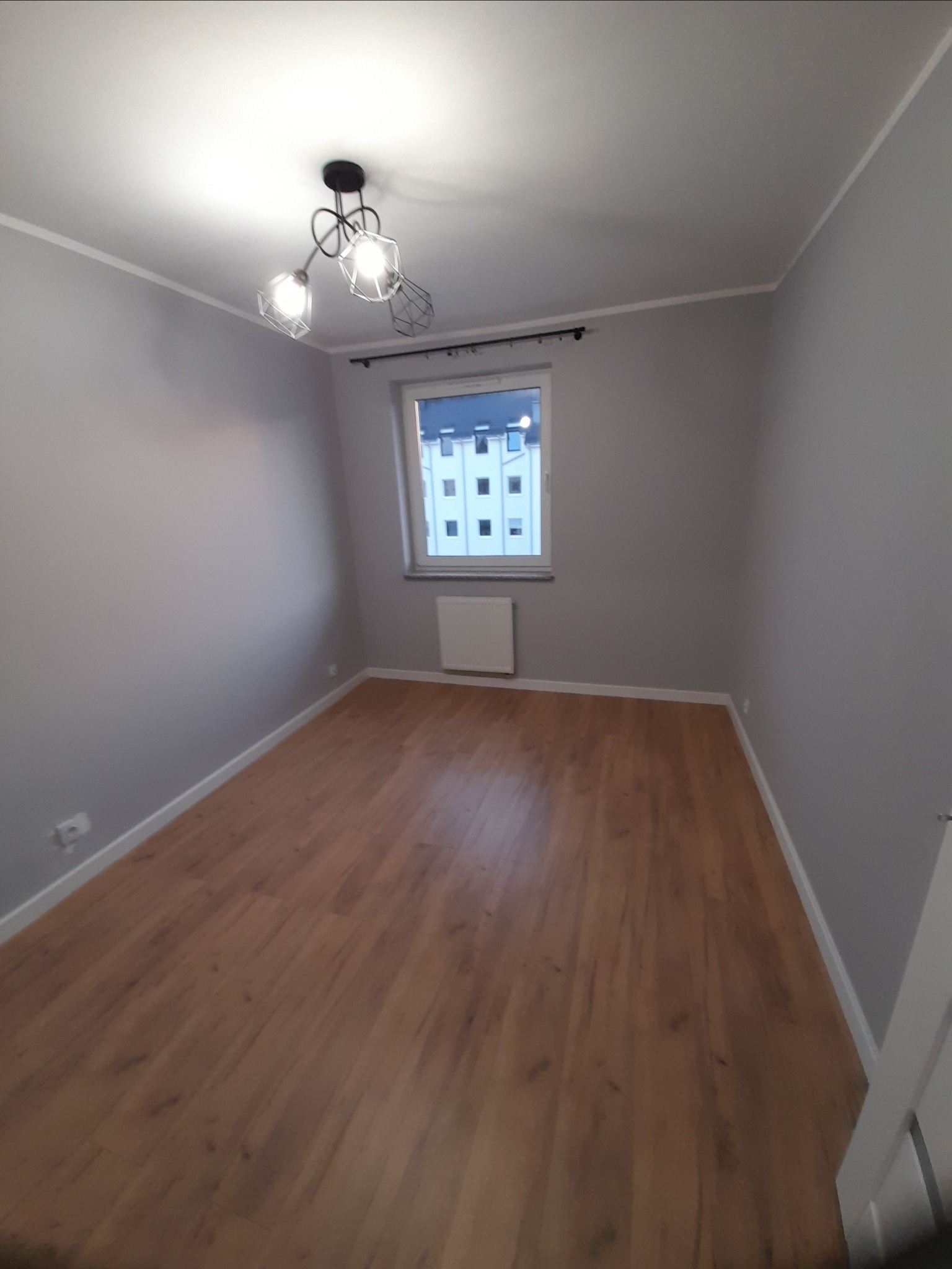 Pomalowane na szaro puste pomieszczenie z panelami podłogowymi, nowoczesną lampą sufitową i oknem z widokiem na budynek.