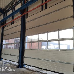 OTWARCI SPÓŁKA Z OGRANICZONĄ ODPOWIEDZIALNOŚCIĄ - Jasna, segmentowa brama przemysłowa z oknami, zamontowana w hali produkcyjnej. Widoczne stalowe konstrukcje nośne i drabina. Brama częściowo otwarta.