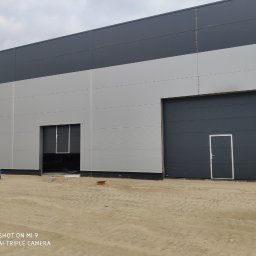 OTWARCI SPÓŁKA Z OGRANICZONĄ ODPOWIEDZIALNOŚCIĄ - Fasada hali przemysłowej z trzema bramami segmentowymi w odcieniach szarości, z czego jedna jest otwarta. Na pierwszym planie teren budowy.