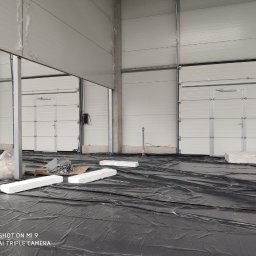 OTWARCI SPÓŁKA Z OGRANICZONĄ ODPOWIEDZIALNOŚCIĄ - Montaż bram przemysłowych segmentowych w hali produkcyjnej. Widoczne białe bramy z drzwiami, czarna folia na podłodze i elementy montażowe.