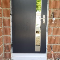 OTWARCI SPÓŁKA Z OGRANICZONĄ ODPOWIEDZIALNOŚCIĄ - Ciemne drzwi zewnętrzne z wąskim przeszkleniem, zamontowane w surowym murze. Widoczne detale montażu i fragment otoczenia z elementami budowlanymi.