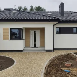 Nowoczesny dom jednorodzinny z białymi ścianami, grafitowym dachem i antracytowymi oknami. Uwagę przykuwają dwuskrzydłowe drzwi wejściowe z długim uchwytem.