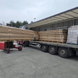 OTWARCI SPÓŁKA Z OGRANICZONĄ ODPOWIEDZIALNOŚCIĄ - Rozładunek pakietów drzwiowych z ciężarówki za pomocą wózka widłowego. Palety zafoliowane, przygotowane do transportu. Widoczne opony ciężarówki i zarys sylwetki osoby.