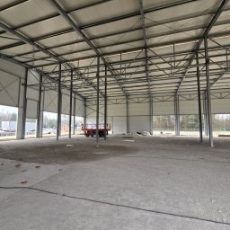 OTWARCI SPÓŁKA Z OGRANICZONĄ ODPOWIEDZIALNOŚCIĄ - Wnętrze hali przemysłowej w trakcie budowy: widoczne stalowe konstrukcje, ściany z paneli, częściowo otwarte bramy i betonowa posadzka. W tle drzewa i częściowo widoczne pojazdy.