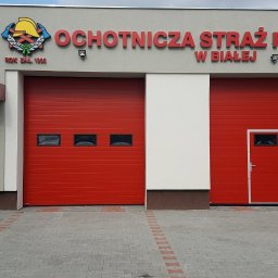 OTWARCI SPÓŁKA Z OGRANICZONĄ ODPOWIEDZIALNOŚCIĄ - Czerwone bramy garażowe segmentowe z oknami i drzwiami w remizie Ochotniczej Straży Pożarnej w Białej, z szarym budynkiem i kostką brukową na pierwszym planie.