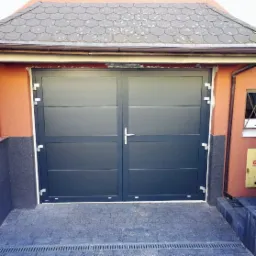 Dwuskrzydłowa, grafitowa brama garażowa z poziomymi panelami, zamontowana w pomarańczowej elewacji z szarym cokołem. Widoczna kostka brukowa przed bramą i dachówka nad nią.