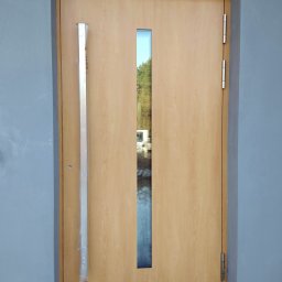 OTWARCI SPÓŁKA Z OGRANICZONĄ ODPOWIEDZIALNOŚCIĄ - Beżowe drzwi zewnętrzne z wąskim przeszkleniem i długim, srebrnym uchwytem w folii ochronnej, zamontowane w szarej ścianie. Widoczne zawiasy i próg.