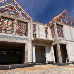 Budowa domu w stanie surowym otwartym: ściany z betonu komórkowego, drewniana konstrukcja dachu, widoczne otwory okienne i drzwiowe na tle błękitnego nieba.