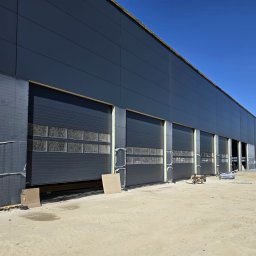 OTWARCI SPÓŁKA Z OGRANICZONĄ ODPOWIEDZIALNOŚCIĄ - Szara hala przemysłowa z segmentowymi bramami garażowymi z oknami, jedna brama częściowo otwarta, widoczne materiały izolacyjne i piaskowe podłoże.