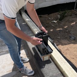 OTWARCI SPÓŁKA Z OGRANICZONĄ ODPOWIEDZIALNOŚCIĄ - Montaż okna aluminiowego: mężczyzna w jeansach ustawia profil okienny na izolacji, przygotowując do instalacji w nowym budynku. Widoczne detale konstrukcyjne.