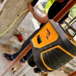 OTWARCI SPÓŁKA Z OGRANICZONĄ ODPOWIEDZIALNOŚCIĄ - Laser krzyżowy Nivel CL2D-G na statywie, używany podczas prac budowlanych. W tle widoczna osoba mierząca coś żółtą taśmą mierniczą.