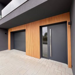 OTWARCI SPÓŁKA Z OGRANICZONĄ ODPOWIEDZIALNOŚCIĄ - Nowoczesna, antracytowa brama garażowa segmentowa i drzwi wejściowe z długim uchwytem, Plewiska. Elewacja z drewnianymi lamelami i szarą kostką.