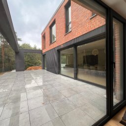 OTWARCI SPÓŁKA Z OGRANICZONĄ ODPOWIEDZIALNOŚCIĄ - Nowoczesny dom z cegły z przesuwnymi oknami aluminiowymi, widok z zewnątrz na taras z szarych płytek. Minimalistyczny design, duże przeszklenia.