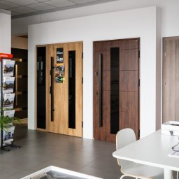 OTWARCI SPÓŁKA Z OGRANICZONĄ ODPOWIEDZIALNOŚCIĄ - Ekspozycja trzech różnych modeli drzwi zewnętrznych w salonie, z widocznymi detalami wykończenia i okuć, uzupełniona o roślinę doniczkową i materiały reklamowe.