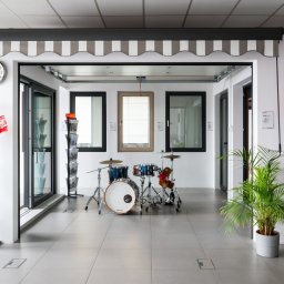 OTWARCI SPÓŁKA Z OGRANICZONĄ ODPOWIEDZIALNOŚCIĄ - Ekspozycja salonowa: markiza w szaro-białe pasy nad oknami, drzwi wejściowe, stojaki z ulotkami i zegar na ścianie, uzupełniona o zestaw perkusyjny i roślinę doniczkową.