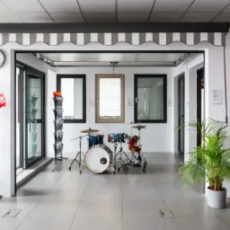 Ekspozycja salonowa: markiza w szaro-białe pasy nad oknami, drzwi wejściowe, stojaki z ulotkami i zegar na ścianie, uzupełniona o zestaw perkusyjny i roślinę doniczkową.