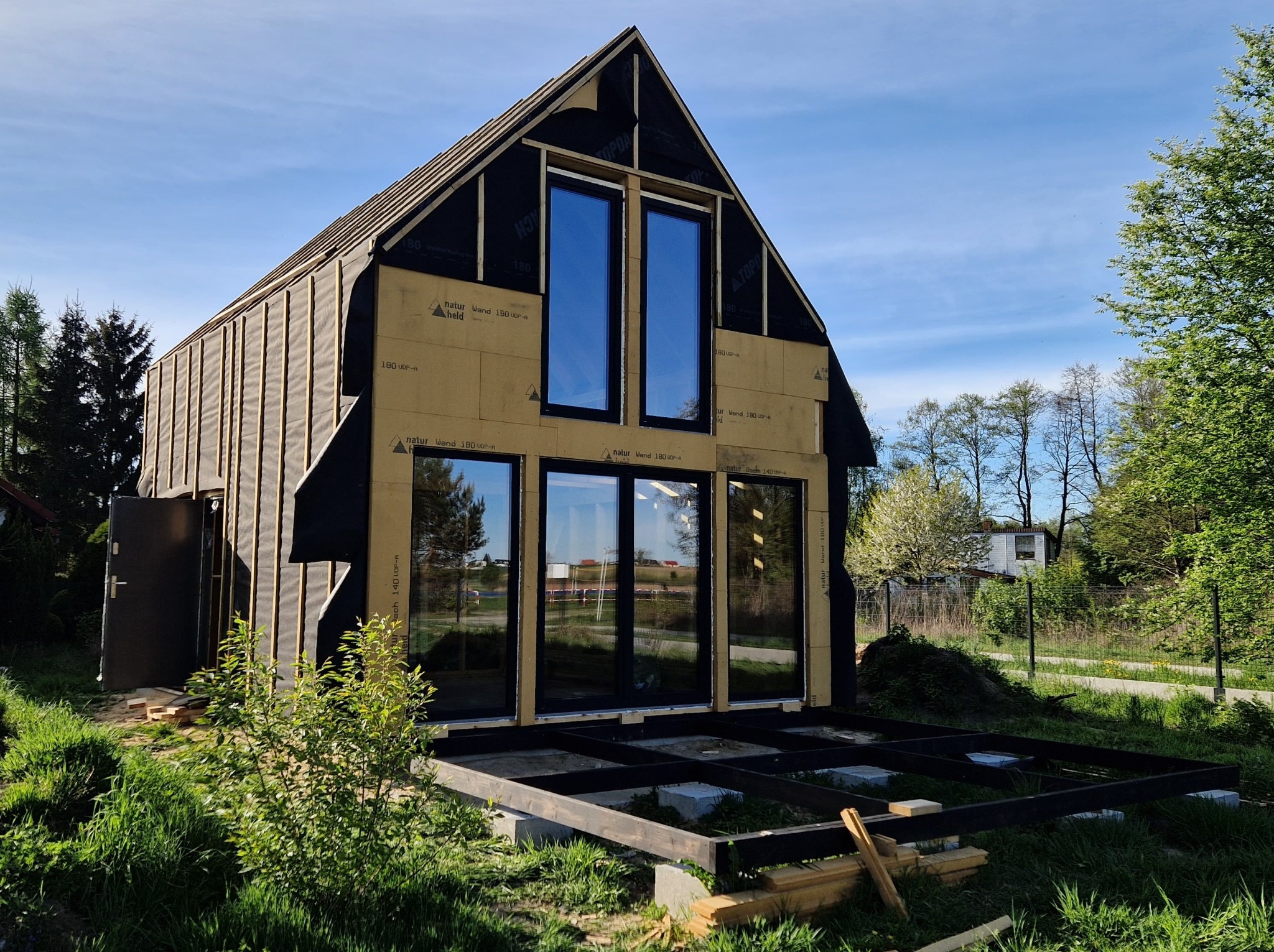 Budowa domu szkieletowego: widoczne okna w czarnych ramach, izolacja ścian, konstrukcja pod taras. Dom w trakcie budowy w otoczeniu zieleni.