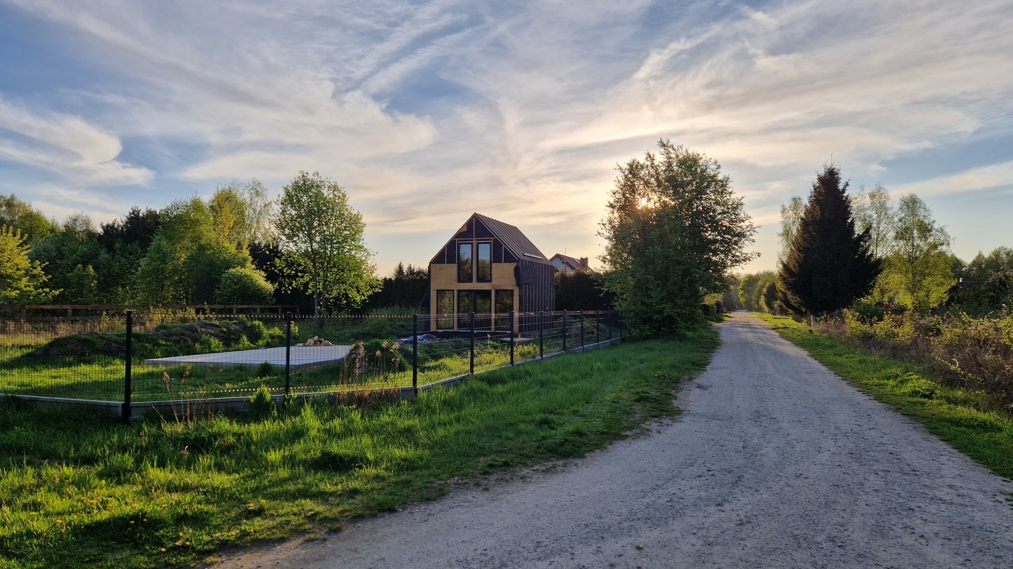 Nowoczesny dom w trakcie budowy, z widocznymi oknami i czarną membraną, otoczony zielenią i ogrodzeniem z siatki, oświetlony ciepłym światłem zachodzącego słońca.