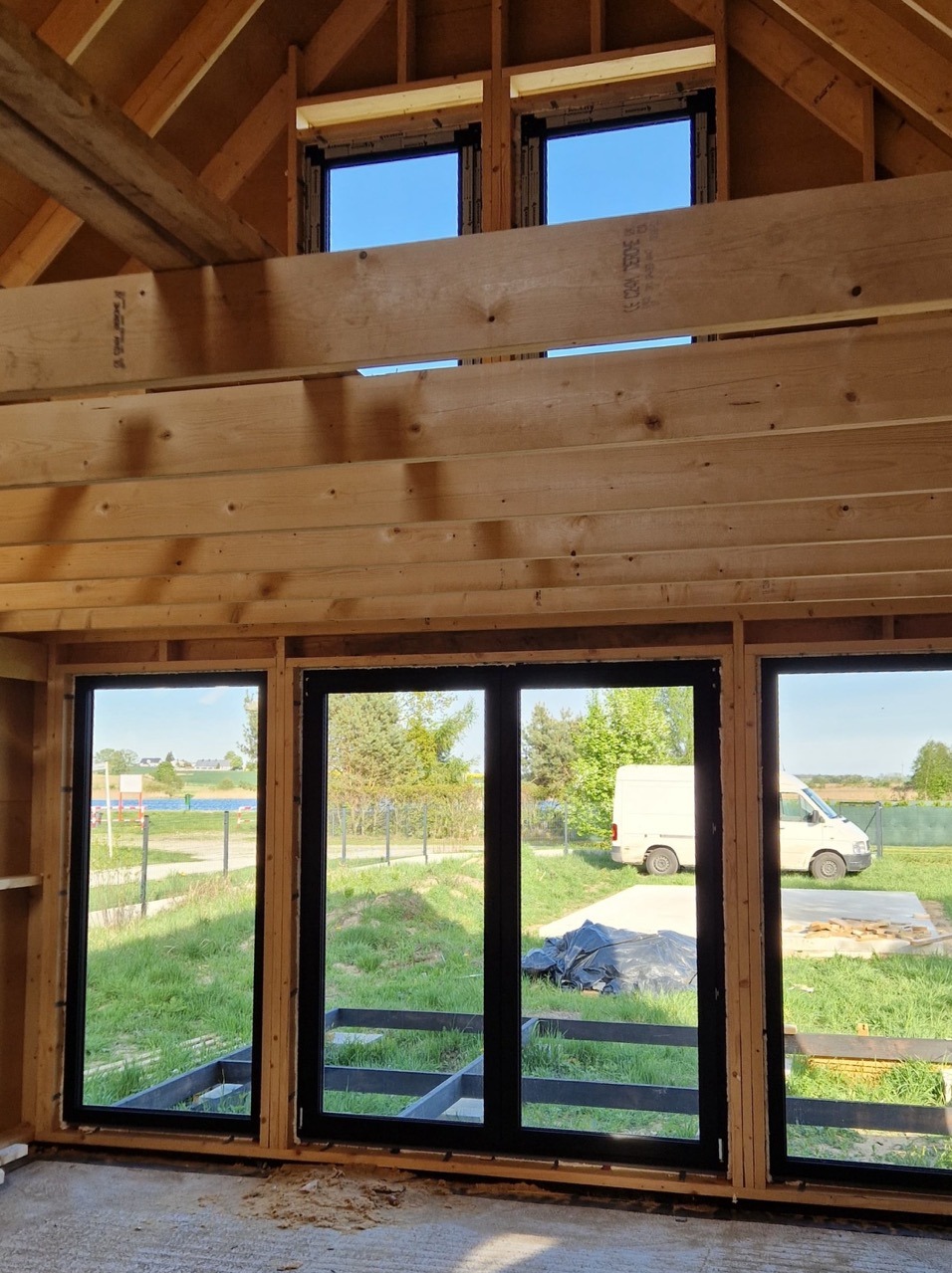 Surowy stan: czarne ramy okienne w drewnianej konstrukcji domu, widok na zewnątrz. Dwa okna dachowe i trzy okna na parterze. Elementy konstrukcyjne w tle.