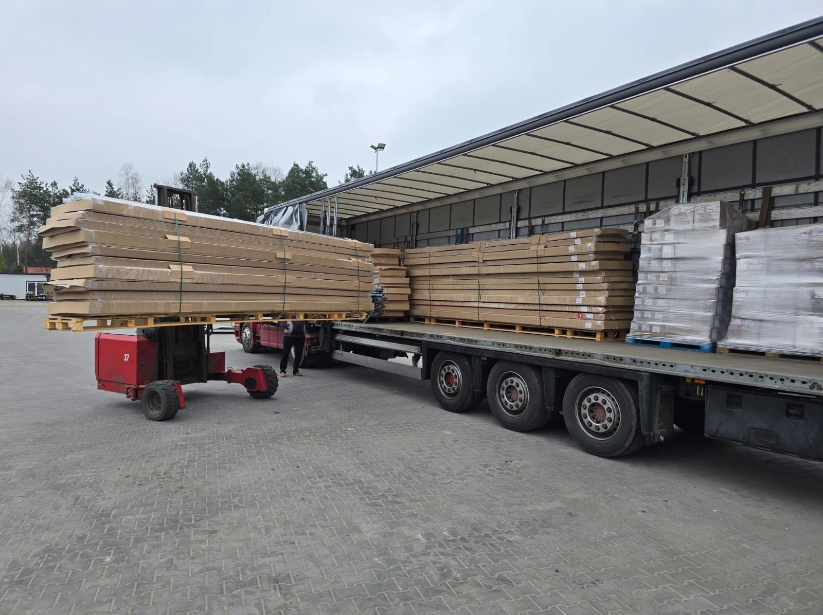 Rozładunek pakietów drzwiowych z ciężarówki za pomocą wózka widłowego. Palety zafoliowane, przygotowane do transportu. Widoczne opony ciężarówki i zarys sylwetki osoby.