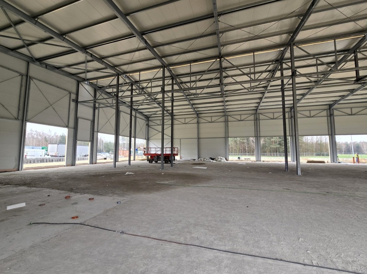 Wnętrze hali przemysłowej w trakcie budowy: widoczne stalowe konstrukcje, ściany z paneli, częściowo otwarte bramy i betonowa posadzka. W tle drzewa i częściowo widoczne pojazdy.