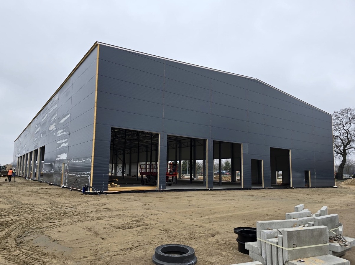 Konstrukcja hali przemysłowej w trakcie montażu, widoczne szare panele ścienne i otwory na bramy, z przodu plac budowy z materiałami.