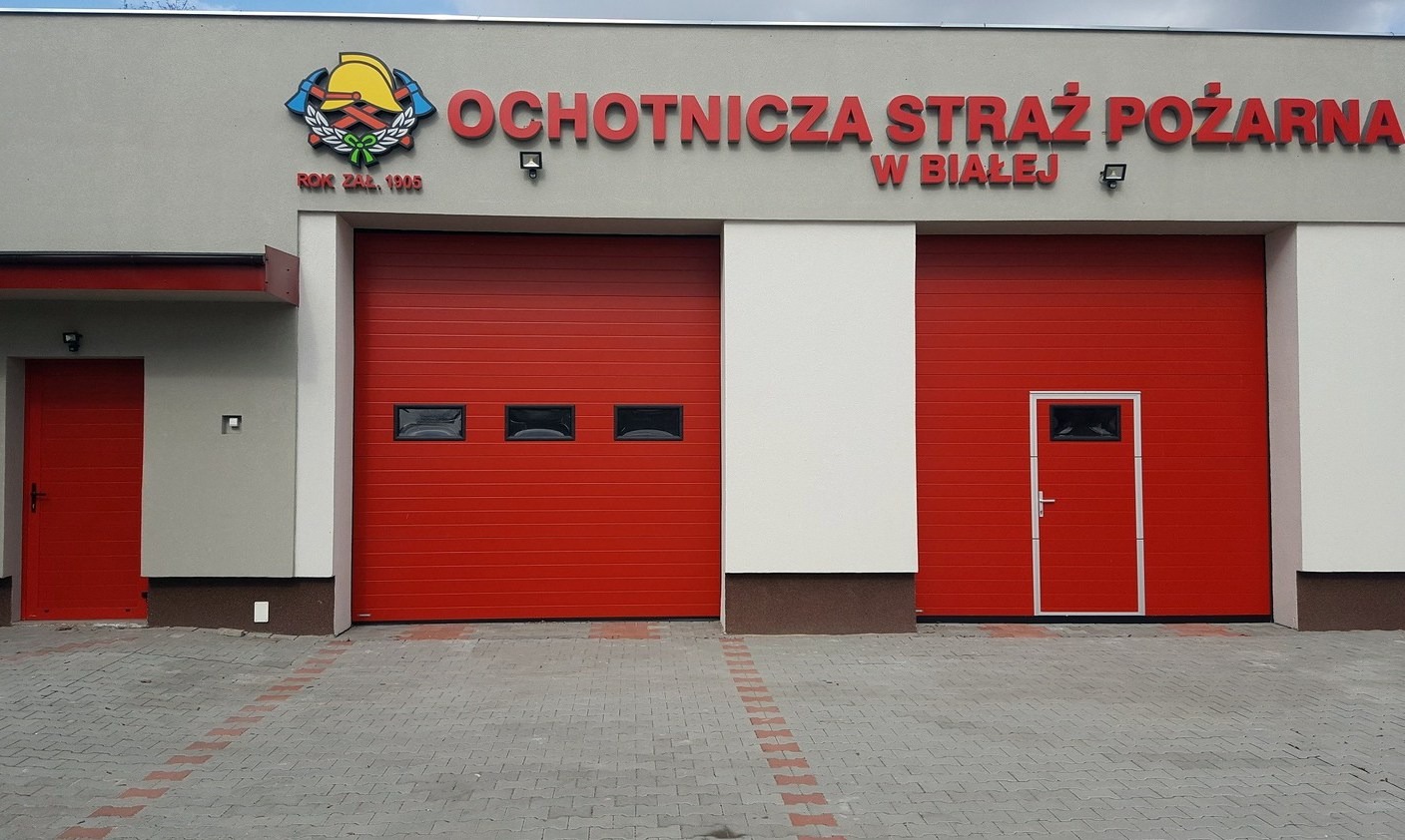 Czerwone bramy garażowe segmentowe z oknami i drzwiami w remizie Ochotniczej Straży Pożarnej w Białej, z szarym budynkiem i kostką brukową na pierwszym planie.
