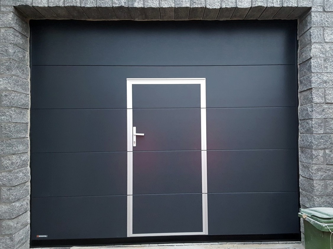 Nowoczesna, antracytowa brama garażowa segmentowa z drzwiami serwisowymi i srebrnym obramowaniem, wkomponowana w kamienną elewację budynku.