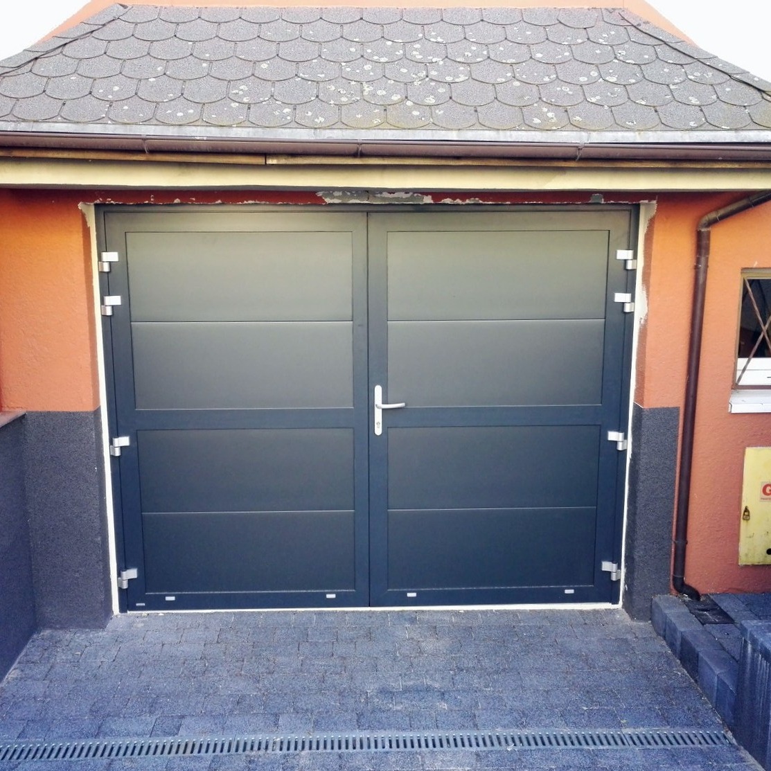 Dwuskrzydłowa, grafitowa brama garażowa z poziomymi panelami, zamontowana w pomarańczowej elewacji z szarym cokołem. Widoczna kostka brukowa przed bramą i dachówka nad nią.