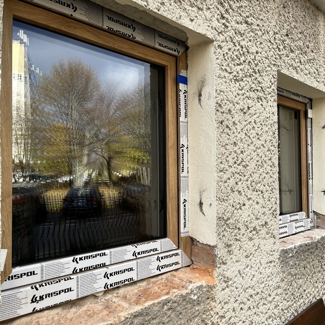Dwa drewnopodobne okna w nierównym, teksturowanym murze. Na oknach zabezpieczenia marki KRISPOL. W szybach odbija się jesienny krajobraz i zabudowa miejska.