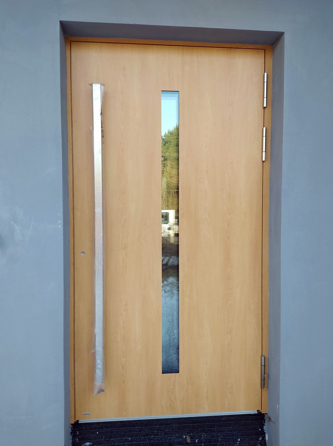 Beżowe drzwi zewnętrzne z wąskim przeszkleniem i długim, srebrnym uchwytem w folii ochronnej, zamontowane w szarej ścianie. Widoczne zawiasy i próg.