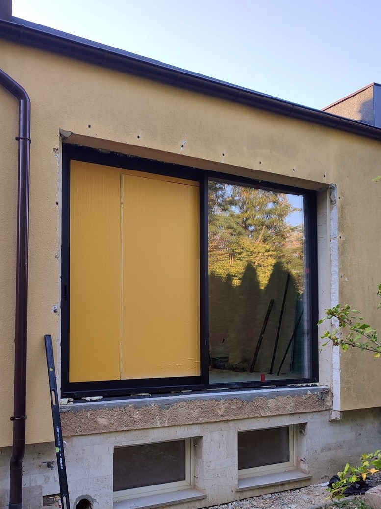 Częściowo zamontowane okno przesuwne z żółtym panelem izolacyjnym po lewej stronie w budynku o żółtej elewacji. Widoczna poziomica.
