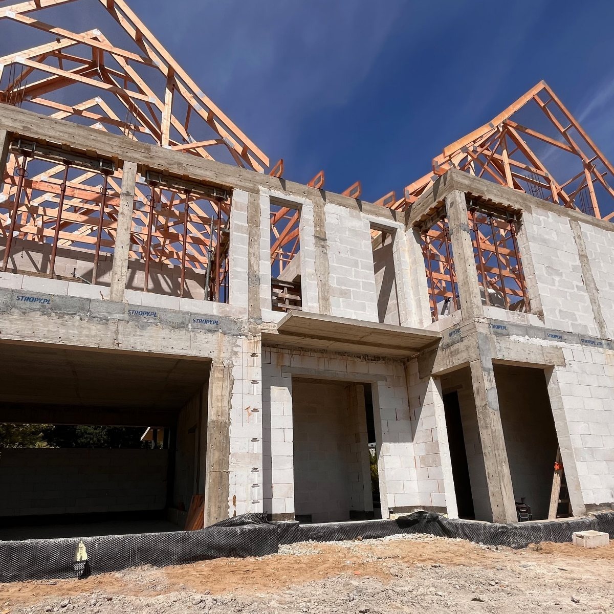 Budowa domu w stanie surowym otwartym: ściany z betonu komórkowego, drewniana konstrukcja dachu, widoczne otwory okienne i drzwiowe na tle błękitnego nieba.