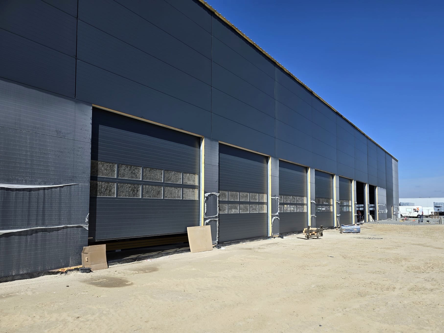 Szara hala przemysłowa z segmentowymi bramami garażowymi z oknami, jedna brama częściowo otwarta, widoczne materiały izolacyjne i piaskowe podłoże.