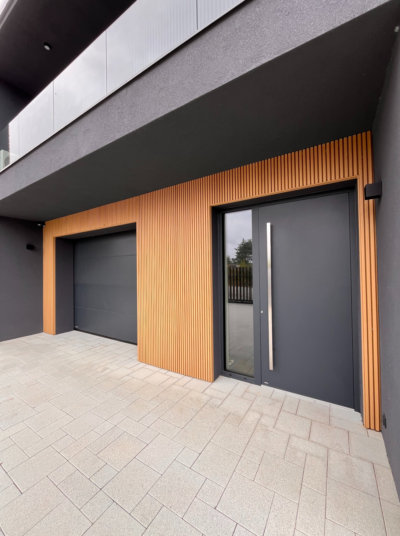 Nowoczesna, antracytowa brama garażowa segmentowa i drzwi wejściowe z długim uchwytem, Plewiska. Elewacja z drewnianymi lamelami i szarą kostką.