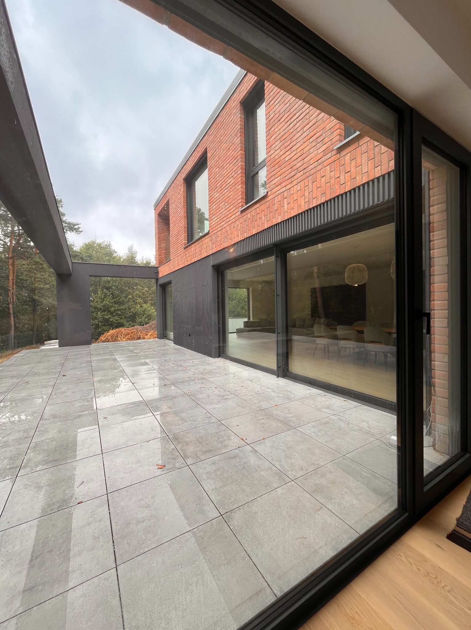 Nowoczesny dom z cegły z przesuwnymi oknami aluminiowymi, widok z zewnątrz na taras z szarych płytek. Minimalistyczny design, duże przeszklenia.
