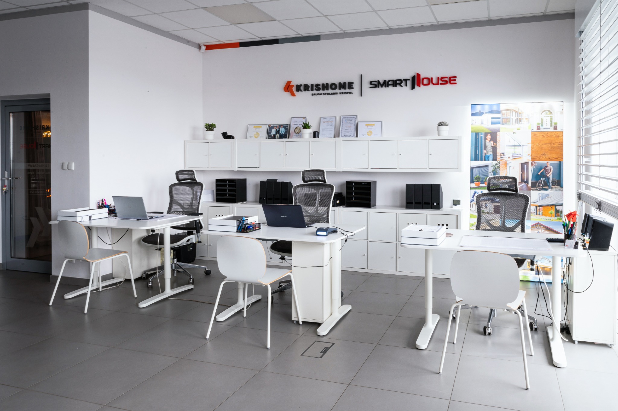 Jasne biuro z białymi meblami i stanowiskami pracy. Na ścianie logo firmy Krishome Smart House i kolaż zdjęć zrealizowanych projektów okien, drzwi i bram.