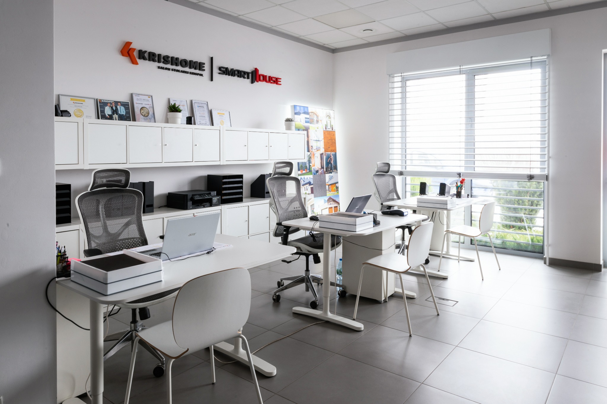 Jasne biuro z białymi biurkami, krzesłami i szafkami. Na ścianie logo firmy 'Krishome Smart House' i certyfikaty. Duże okno z żaluzjami wpuszcza naturalne światło.